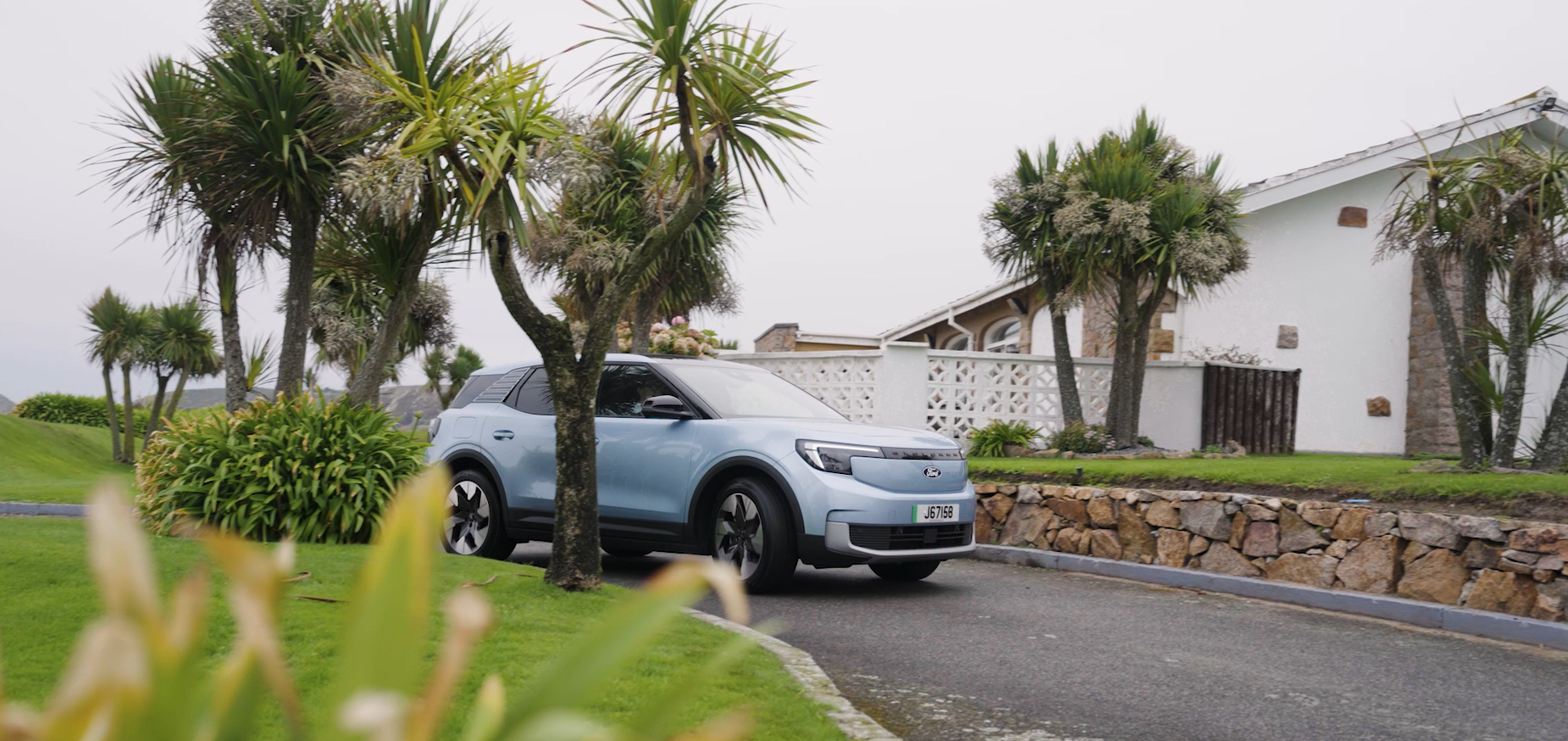 A light blue electice Ford Explorer pulling into a drivewhich has grass and palm trees either side. A house sits in the backgroud