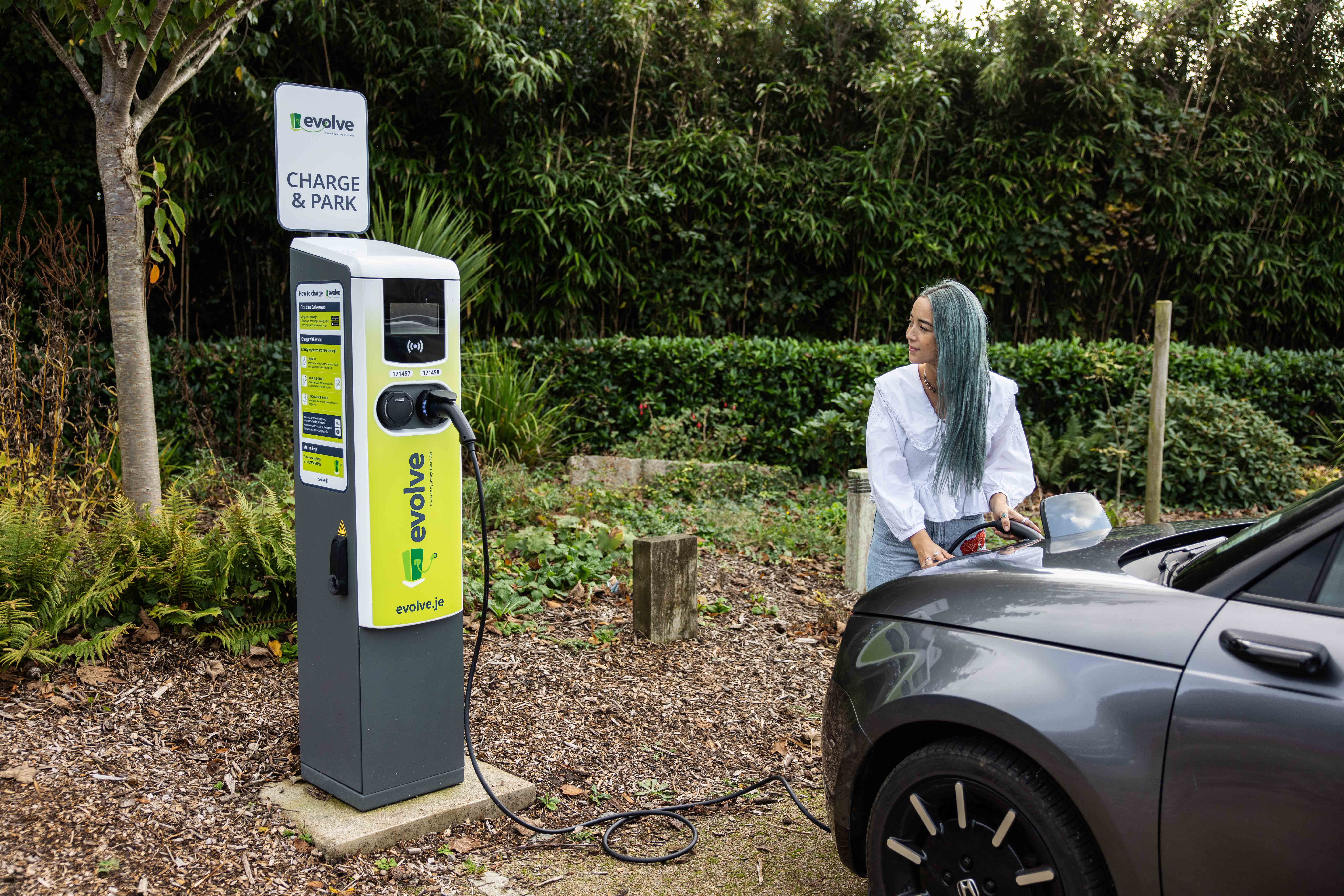 A person with blue-tinted hair connects a charging cable from an EV charger to a dark gray electric car.